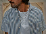 A man in Blue de Gênes BdG S/S Grandad - Snow White shirt, featuring corozo nut buttons, layers it over a white cotton slub yarn henley. He wears a silver chain necklace and has a mustache, standing in front of blurred neutral-toned shapes.