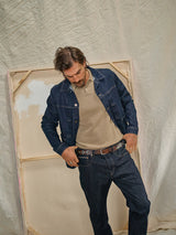 A man in a Blue de Gênes Cosimo Trucker Jacket - Dark Blue Denim and jeans, paired with a beige sweater, stands before a large canvas. He gazes downward with one hand in his pocket against a backdrop of rumpled, light-colored fabric.