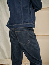 A person poses sideways in a Cosimo Trucker Jacket - Dark Blue Denim by Blue de Gênes, paired with dark denim jeans. The jacket’s visible stitching and casual fit enhance the relaxed and stylish vibe against a soft, neutral background.
