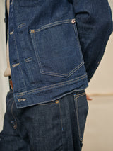 Close-up of someone in a Blue de Gênes Cosimo Trucker Jacket - Dark Blue Denim and jeans, showcasing visible stitching and a large front pocket. With hands behind their back, part of a beige shirt is visible underneath.
