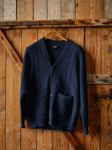 The Achille Cardigan - Navy by Blue de Gênes, featuring Corozo button closure and two front pockets, hangs on a wooden hanger against a rustic wooden door.