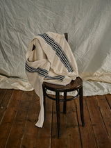 The Cappa Stripe Cardigan - Whitecap by Blue de Gênes, featuring navy stripes on cream-colored cotton yarn, is draped over a dark wooden chair on a wooden floor, set against a crumpled off-white fabric backdrop.