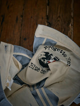 A pair of open Palto Riga Chino - Blue Stripe trousers by Blue de Gênes on a wooden floor, featuring a button fly. The interior label includes an illustration, while the cotton fabric features a striped pattern lining for a relaxed fit.