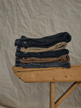 A stack of Blue de GĂȘnes Paulo Pavia 1 Chino pants, including jet black, made in Italy with premium stretch cotton, rests on a wooden table against a draped grayish fabric backdrop.