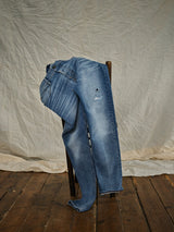 The Blue de Gênes Vinci Super Used Jeans - Used, featuring patches and slight fading, expertly crafted in Italy, are draped over a wooden chair against a light beige fabric backdrop on a wooden floor.