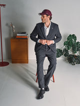 A man in a dark suit and maroon cap sits on a stool, showcasing Blue de Gênes Kyoto Pant - Black Pattern beside a wooden cabinet, red coat rack, books, a plant, and a white wall.