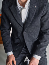A person in a Blue de Gênes Fukui Blazer - Black Pattern with relaxed tailoring and a small lapel pin, over a white dress shirt, is kneeling with their face partially out of frame.