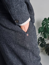 A person wears Blue de Gênes Kyoto Pant in Black Pattern with a white shirt cuff showing and a hand in the pocket; a green monstera plant appears blurred in the background.