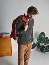 A man indoors drapes a rust jacket over his shoulder, wearing the Blue de Gênes Malakka Polo Knit in Army and brown pants. A wooden cabinet with books and a green houseplant is in the background.