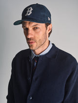 A man stands against a plain light background wearing the Blue de Gênes Gifu Knit Cardigan in navy, along with a baseball cap featuring a B logo, a blue striped shirt, and a tie.