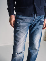 A person wearing a dark sweater and Blue de Gênes Giacomo Osaka Jeans in dark denim, featuring a relaxed, loose fit with visible patches and rips, stands indoors against a plain light-colored background.