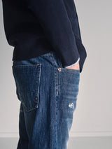 Wearing Blue de Gênes Giacomo Osaka Jeans in Dark Denim, a person faces sideways against a plain light background, one hand in their pocket and dressed in a navy blue sweater.