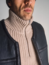 Close-up of a man wearing the Blue de Gênes Yomaha Halfzip Knit - Fungi, a beige chunky knit sweater with a front zipper, paired with a black leather vest. Only his lower face and torso are shown.