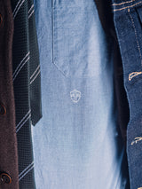 Close-up of a person wearing a blue shirt with a white embroidered shield, Nekutaki Tie #D - Green Stripe by Blue de Gênes, brown coat, and denim jacket. Only the chest area is shown.
