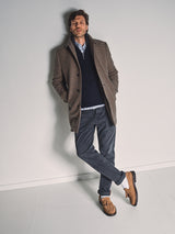 A man stands against a plain wall wearing the Blue de Gênes Massone Check Coat in Multi Check, paired with a navy sweater, light blue shirt, grey pants, and tan loafers. One hand is in his pocket and his legs are crossed at the ankles.