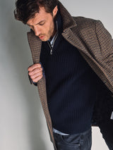 A man with tousled hair stands against a plain background, wearing the Blue de Gênes Yomaha Halfzip Knit in navy under a brown houndstooth coat. He looks down, holding his coat open with one hand.