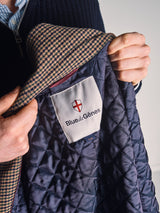 A person holds open the Massone Check Coat - Multi Check by Blue de Gênes, revealing the inside label with the Blue de Gênes name and a red and white shield logo. The coat features a houndstooth pattern on the collar.