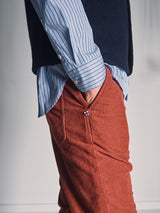 A person stands with one hand in the pocket of Blue de Gênes BGpaulo Ottawa Trousers in Henna, paired with a blue and white striped button-up shirt and a dark vest, against a softly lit plain background.