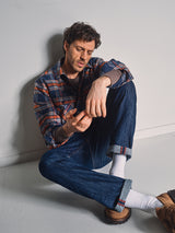 A man with wavy hair sits on the floor against a white wall, looking down thoughtfully. He wears a relaxed fit Blue de Gênes Yodo Check Overshirt - Multi Check, jeans, white socks, and brown shoes.