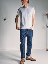 A man stands indoors wearing Blue de Gênes Giacomo Dark Horse Jeans in Denim Blue, a white t-shirt, and brown shoes. One hand is in his pocket as he looks to the side against a plain, minimally decorated background.