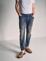 A person stands against a plain background wearing a white t-shirt, Blue de Gênes Repi Dark Vintage Jeans in mid blue denim, and brown suede loafers, hands in pockets. Their head is not visible in the image.