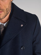 Close-up of a man in the Blue de Gênes Shiba Melton Coat in dark navy, paired with a white shirt and patterned tie. A pin with a red cross on white decorates the lapel, and only the lower half of his face is visible.