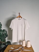 A BGoscar T-Shirt - Snow Fall by Blue de Gênes in regular fit hangs on a wooden hanger against a light gray wall. In front, a wooden table holds spools of thread, measuring tape, tags, safety pins, and a leafy plant sits on the left.