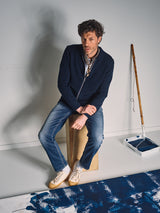 A man in a navy jacket, jeans, and the Blue de Gênes Siruku Scarf - Mixed Colours sits on a wooden stool beside a paint roller, tray of dark blue paint, and a partly painted canvas. The softly lit background is plain.