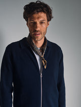 A man with wavy brown hair wears a dark zip-up jacket, white shirt, and the Blue de Gênes Siruku Scarf in mixed colours tied around his neck, standing against a plain gray background.