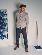 A man wearing the Blue de Gênes Sakura Knit Cardigan in Stone, dark jeans, and brown shoes stands indoors by a blue paint roller tray and a canvas splattered with blue.