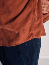A person wears the Blue de Gênes Zamboni Velu Shirt in Henna, a rust-toned baby corduroy shirt with a small shield logo, paired with dark blue Blue de Gênes corduroy pants and stands with one hand partially visible at their side.