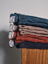 Four pairs of Blue de Gênes BGpaulo Ottawa Trousers in blue, beige, red, and navy are stacked on wood. Made in Italy from dobby corduroy, each features visible buttons and branded waistband labels.