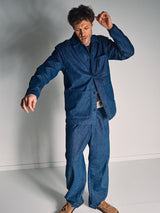 A man stands indoors in mid-motion, wearing the Blue de GĂȘnes Shogun Fano Pant in Denim Blue with a loose-fitting denim jacket and light brown shoes.