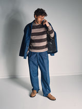 A man stands in a minimal studio wearing the Blue de Gênes Yuma Fano Jacket in Denim Blue, paired with blue denim pants, a brown and gray striped sweater, and tan shoes, looking down with one hand touching his face.
