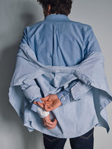 A person seen from behind wears the Sandro Oxford Shirt in blue by Blue de Gênes over a striped shirt, hands crossed behind their back and fingers holding the striped layer, against a plain gray background.