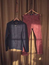 A navy blue button-up cardigan is paired with Blue de Gênes Paulo Tucan Chino - Chipotle, crafted from fantasy comfort stretch corduroy. Both garments hang on wooden hangers against a brown curtain, with soft sunlight casting subtle shadows.