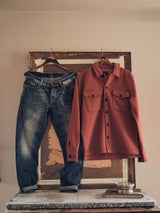 The Pampa Overshirt - Ochre Red by Blue de GĂȘnes, featuring Corozo buttons and two front pockets, hangs on a hanger next to blue jeans against a textured brown wall. Below, a small metal bowl rests on a ledge, enhancing the rustic, vintage aesthetic.