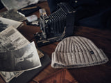 A vintage camera rests on a wooden table with old black and white photos. Nearby, a Blue de Gênes Oscar Melange Beanie in Pebble Beige, made from soft wool blend, adds texture alongside a leather-bound book and various small objects.