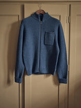 The Aimone Knit Cardigan by Blue de GĂȘnes, in a textured coarse pearl knit style and sugar blue hue, features ribbed cuffs and hem with a small left chest pocket. It hangs on a wooden hanger against a beige wardrobe.