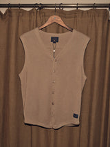 The Zorba Knit Waistcoat - Taupe by Blue de Gênes, a beige sleeveless vest made from soft merino wool with corozo nut buttons, hangs on a wooden hanger against matching beige curtains. A label near the bottom edge enhances its elegant simplicity.