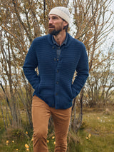 A bearded man in a blue knit cardigan, Paulo Tucan Chino - Biscotto by Blue de Gênes, and a white beanie stands with hands in pockets on grassy terrain with bare trees behind.