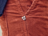 A close-up of the Chipotle Paulo Tucan Chino by Blue de Gênes, showcases red-orange fantasy comfort stretch corduroy with visible vertical lines. A metal pin shaped like a shield with a white cross marks its luxurious made in Italy craftsmanship.