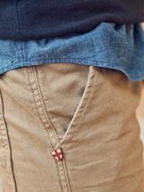 A close-up shows a person in khaki pants featuring a red and white shield emblem near the pocket, paired with a Miguel Chambray Shirt in Lt. Denim Blue by Blue de Gênes and a dark sweater, all crafted from premium cotton fabric for a crisp, timeless style.