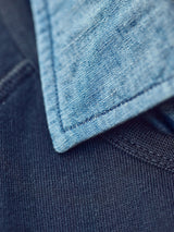 A close-up of the Miguel Chambray Shirt in Lt. Denim Blue by Blue de Gênes shows its visible stitching contrasting beautifully with a dark blue knit, highlighting the interplay between these cotton fabrics.