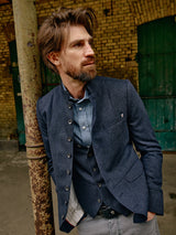 A bearded individual stands outside by a rusted pole, donning the Blue de Gênes Canna Santo Gilet in navy over a denim shirt and gray pants. They gaze sideways against a backdrop of brick walls and green doors.