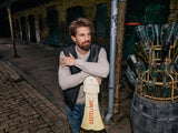 A bearded man leans on a large champagne bottle sculpture in a dimly lit, industrial setting. Wearing an Adamo Knit - Pebble Beige by Blue de Gênes, he stands next to a barrel with upside-down bottles. The brick wall backdrop adds rustic charm.