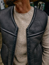 Close-up of a person wearing the Adamo Knit - Pebble Beige by Blue de Gênes under a black leather vest. Soft lighting accentuates the textured mélange yarn. The focus is on the clothing, not the persons face.