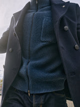 A person wears a dark navy double-breasted coat with prominent buttons over a cozy Blue de GĂȘnes Aimone Knit Cardigan in Sugar Blue, standing outdoors on a cloudy day, paired with dark jeans.