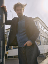 A bearded man in a Blue de GĂȘnes Aimone Knit Cardigan - Sugar Blue and lambswool sweater stands outdoors, leaning against a pole. His windswept hair enhances his rugged charm as he gazes at the camera, with a sunlit building in the background.