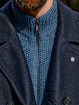 A close-up features a person in a navy coat with an emblem of a red cross on a white shield and black buttons, worn over the Aimone Knit Cardigan - Sugar Blue by Blue de GĂȘnes. Their lower face is partially visible, framed by their beard.